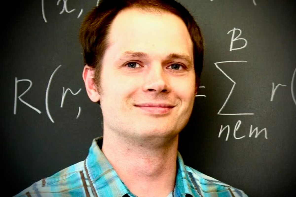 A man in a collared plaid shirt smiles, standing in front of a chalkboard with forumulas.